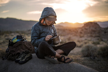 Happy female hiker eating food while sitting on sand dune - CAVF18271