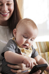 Glückliche Mutter, die ihr Baby hält, während sie zu Hause ein Mobiltelefon benutzt - CAVF18522