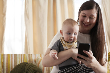 Happy mother showing mobile phone to baby while sitting on sofa at home - CAVF18524