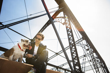 Niedriger Blickwinkel auf einen Mann, der mit einer Bulldogge auf einer Stützmauer gegen den Himmel sitzt - CAVF18754