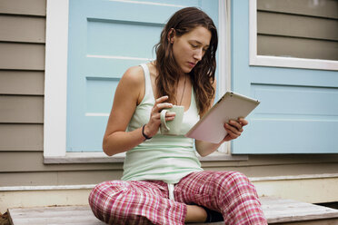 Junge Frau, die einen Tablet-Computer vor dem Haus benutzt und einen Kaffeebecher hält - CAVF18879