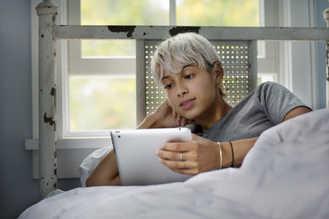 Junge Frau schaut auf einen Tablet-Computer, während sie sich auf dem Bett entspannt, lizenzfreies Stockfoto