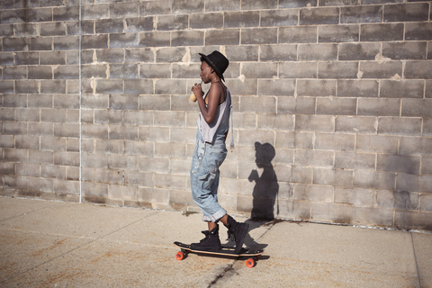 Seitenansicht einer Frau, die einen Smoothie trinkt und auf einem Skateboard steht, lizenzfreies Stockfoto