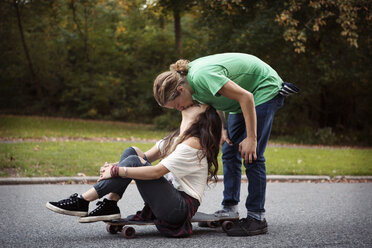 Man kissing woman sitting on skateboard at street - CAVF19010