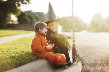 Fröhliche Geschwister für Halloween-Party gekleidet sitzen an der Straße - CAVF19059