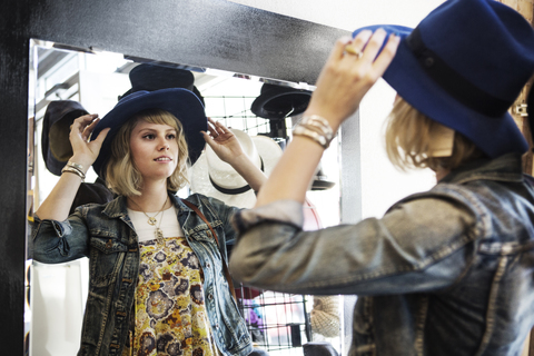 Frau probiert einen Hut an, während sie vor einem Spiegel steht, lizenzfreies Stockfoto