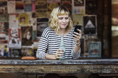 Frau, die ein Smartphone benutzt, während sie eine Limonadenflasche in einem Cafe hält, lizenzfreies Stockfoto