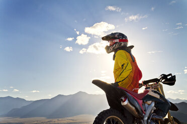 Biker looking at mountains while standing by dirt bike against sky - CAVF19356