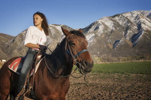 Weiblicher Teenager, der wegschaut, während er auf einem Pferd vor einem Berg sitzt - CAVF19430
