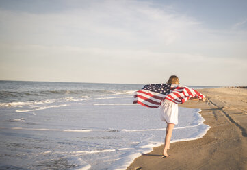 Rückansicht eines jungen Mädchens, das eine amerikanische Flagge hält, während es am Strand gegen den Himmel läuft - CAVF19662