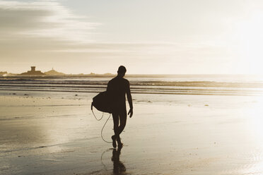 Silhouette eines Mannes, der ein Surfbrett trägt, während er bei Sonnenuntergang am Strand spazieren geht - CAVF19686