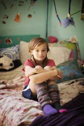 Portrait of confident girl hugging knees while sitting on bed in room - CAVF19728