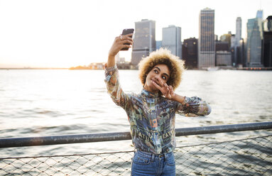 Woman taking selfie through mobile phone while standing against river - CAVF19767