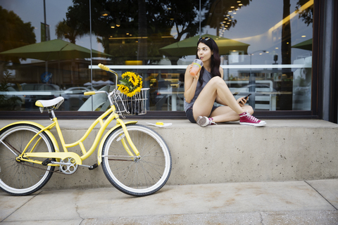 Schöne Frau trinkt Saft, während sie auf einer Stützmauer sitzt und Fahrrad fährt, lizenzfreies Stockfoto