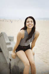 Cheerful woman sitting on bench at beach - CAVF19798