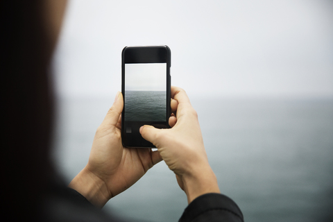 Ausgeschnittenes Bild einer Frau, die mit ihrem Mobiltelefon das Meer fotografiert, lizenzfreies Stockfoto