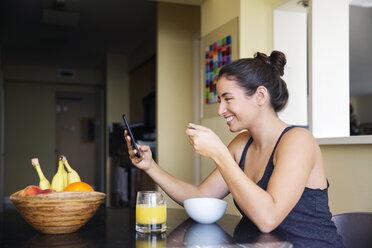 Smiling woman using smart phone while eating breakfast on table at home - CAVF19840