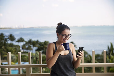 Glückliche Frau, die ihr Smartphone benutzt, während sie Kaffee gegen das Meer trinkt - CAVF19848