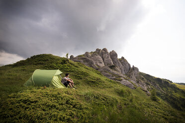 Low angle view of hiker sitting by tent on mountain against cloudy sky - CAVF19906