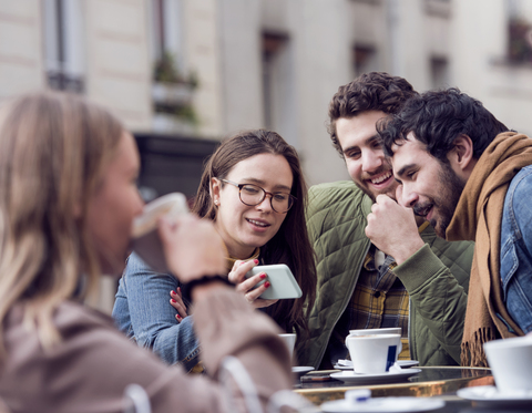 Glückliche Frau zeigt männlichen Freunden in einem Straßencafé ihr Smartphone, lizenzfreies Stockfoto