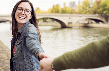 Cheerful young woman holding man's hand by river - CAVF19936