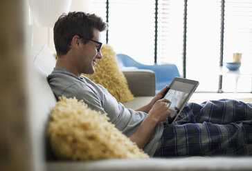 Smiling man using tablet while resting on sofa at home - CAVF19984