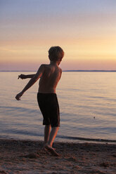 Rückansicht eines Jungen, der am Strand einen Stein ins Wasser wirft - CAVF20008