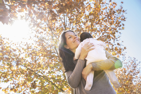 Lächelnde Mutter, die ein Kleinkind trägt, während sie an einem sonnigen Tag an einem Baum steht, lizenzfreies Stockfoto