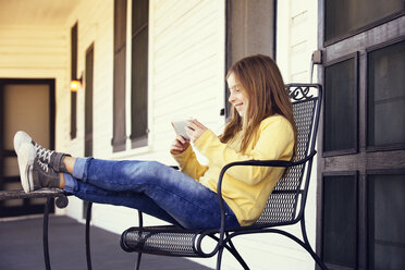 Happy girl using smart phone while sitting on chair - CAVF20062