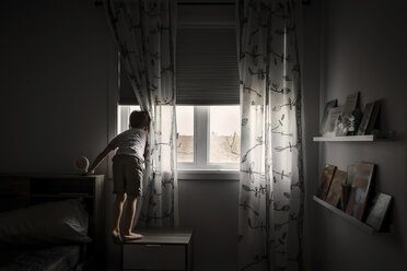 Rückansicht eines Jungen, der durch ein Fenster schaut, während er zu Hause auf einem Schrank steht - CAVF20182