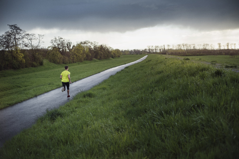 Rückansicht eines männlichen Sportlers, der auf der Straße inmitten eines grasbewachsenen Feldes läuft, lizenzfreies Stockfoto