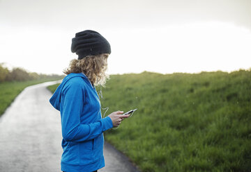 Seitenansicht weibliche Athletin mit Smartphone auf der Straße inmitten grasbewachsenen Feld gegen klaren Himmel - CAVF20200