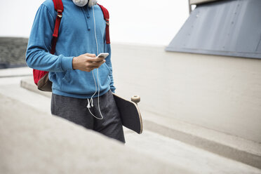 Mittelteil eines Mannes, der ein Mobiltelefon benutzt, während er ein Skateboard auf einem Parkplatz trägt - CAVF20234