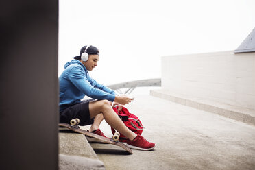 Man listening music while sitting on footpath at parking lot - CAVF20236