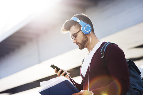 Junger Mann, der ein Telefon benutzt, während er Musik gegen eine Brücke hört - CAVF20286