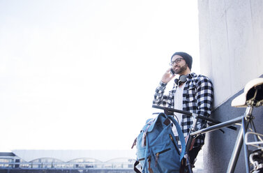 Young man talking on phone while leaning on wall by bicycle against clear sky - CAVF20292