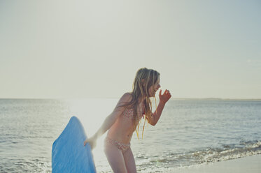 Fröhliches Mädchen hält Surfbrett am Strand gegen den Himmel - CAVF20415