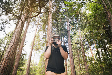 Mann, der durch die Kamera fotografiert, während er gegen Bäume im Wald steht - CAVF20534