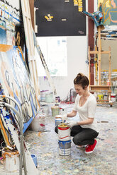 Glückliche Künstlerin, die sich hockt, während sie den Pinsel in eine Farbdose im Kunststudio taucht - CAVF20811