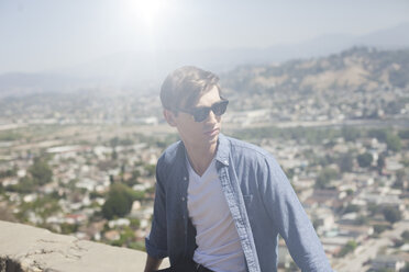 Young man sitting, overlooking city on sunny day - CAVF20864