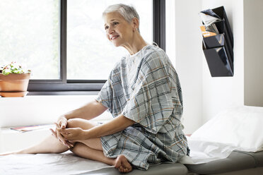 Glückliche, nachdenkliche Frau, die wegschaut, während sie in der Klinik auf dem Bett sitzt - CAVF21046