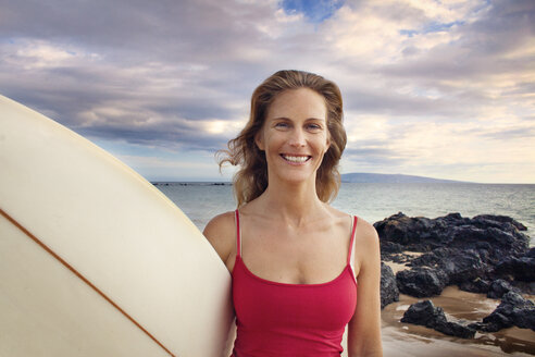 Porträt einer lächelnden Frau mit Surfbrett am Meeresufer gegen bewölkten Himmel - CAVF21985