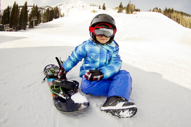 Mädchen mit Skibrille sitzt mit Snowboard auf einem verschneiten Feld - CAVF22021