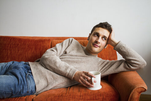 Nachdenklicher Mann hält Kaffeetasse, während er zu Hause auf dem Sofa liegt - CAVF22064