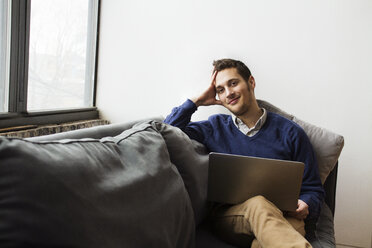 Smiling man using laptop while sitting on sofa at home - CAVF22067