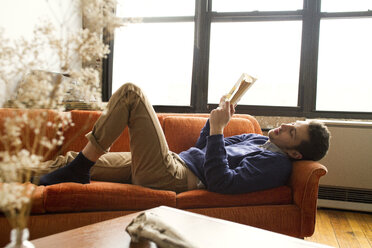 Man reading book while lying on sofa at home - CAVF22069