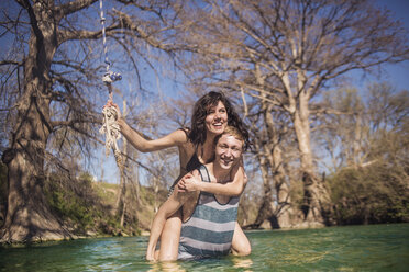 Porträt einer fröhlichen Freundin, die ihren Freund im Fluss vor kahlen Bäumen an einem sonnigen Tag huckepack nimmt - CAVF22132