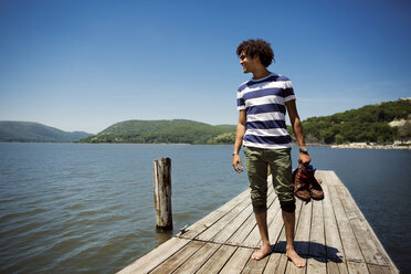 Smiling man holding shoes while standing on jetty over lake - CAVF22215