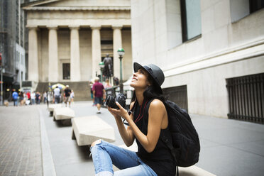 Woman with camera carrying backpack sitting on bench in city - CAVF22245