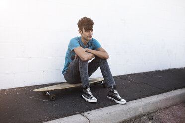 Thoughtful man sitting on skateboard on sidewalk against white wall - CAVF22259